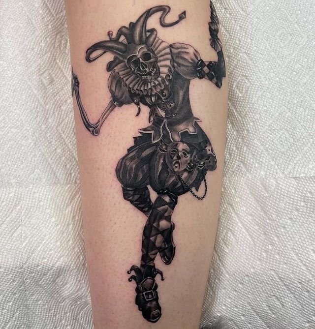 Black-and-grey illustrative skeleton jester leg tattoo